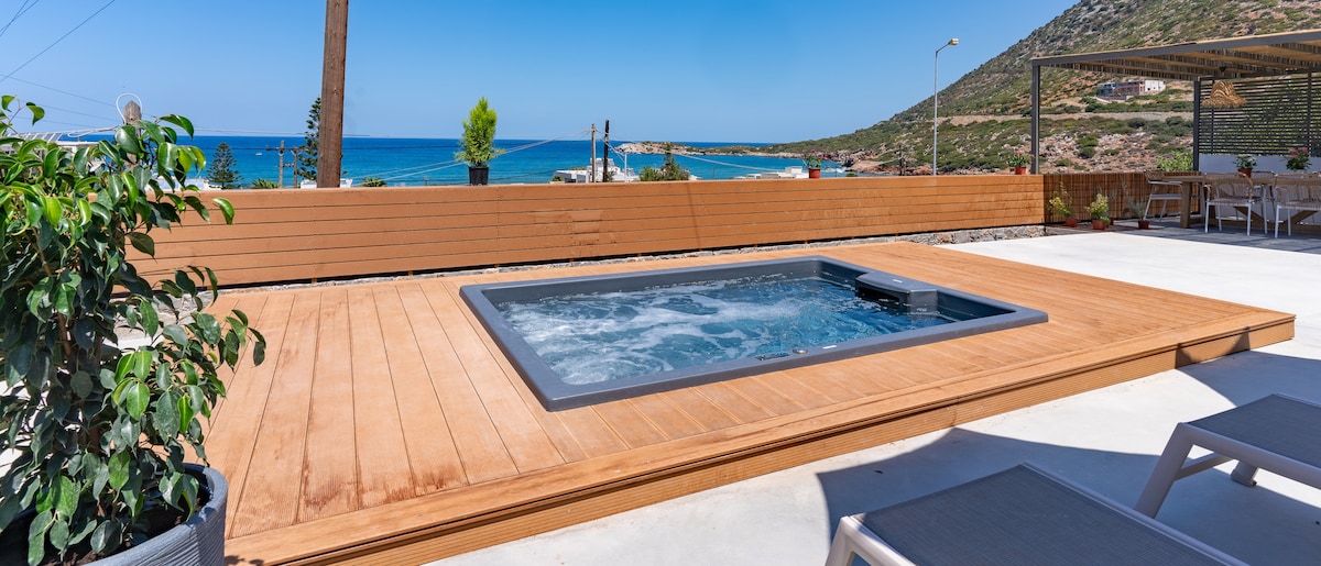 A wooden deck surrounds a hot tub, offering ocean views in the background. The area is designed with light-colored loungers and a small green plant, creating a comfortable space for relaxation.