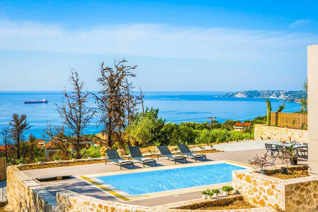 A private pool is set against a backdrop of the sea, featuring several lounge chairs positioned nearby. The landscape includes varied vegetation and stone pathways leading towards the water's edge, with distant views of the coastline and hills under a clear blue sky.