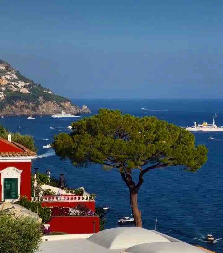 A coastal view captures a vibrant blue sea, with boats gently bobbing in the water. A prominent green tree stands on a hillside, providing a contrasting element to the surrounding architecture, which features a red exterior and white trim. The backdrop showcases a mountainous coastline.