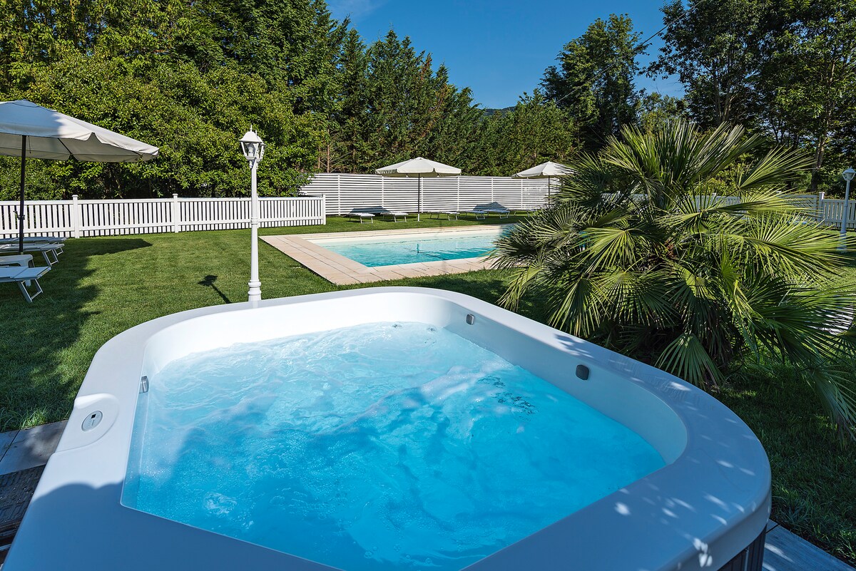A white outdoor hot tub is positioned in the foreground, featuring a calming blue water surface. In the background, a swimming pool is visible, surrounded by sun loungers and shaded by umbrellas. Lush green foliage complements the serene outdoor setting.