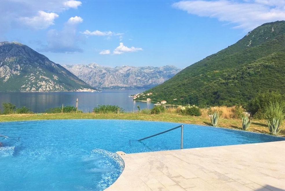 A tranquil infinity pool is set against the backdrop of the Bay of Kotor, with rolling green hills and distant mountains extending towards the horizon. The clear blue water blends seamlessly into the sky, creating a peaceful ambiance.