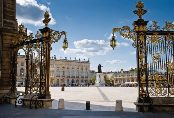 Ornate golden gates frame a vast square under a clear sky. In the center, a statue stands prominently, surrounded by elegant buildings with architectural details. The expanse of the square is visible, creating a serene atmosphere for leisurely strolls.