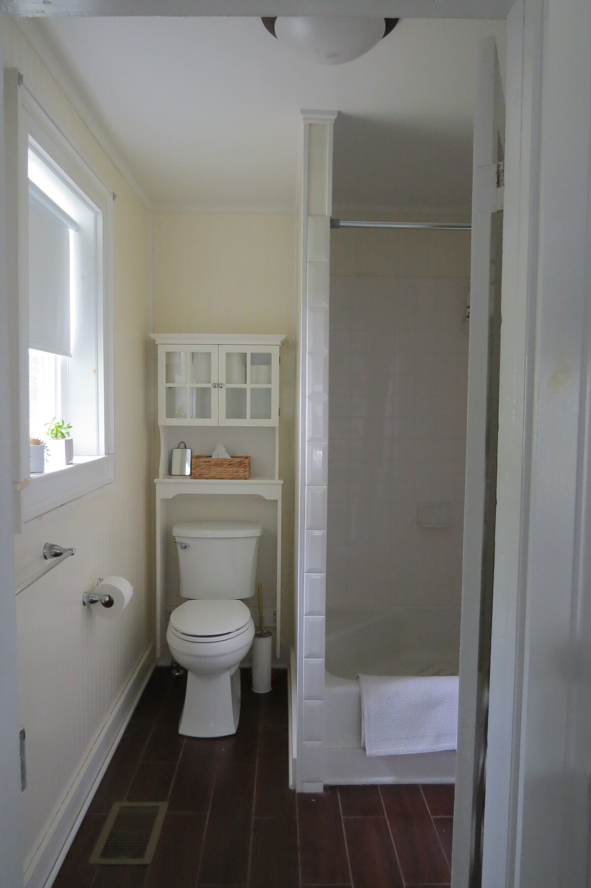 A clean bathroom design features a white shower enclosure and a spacious bathtub. A toilet is positioned beside a small shelving unit, holding toiletries. Light enters through a window, and warm wood flooring adds a touch of warmth to the space.