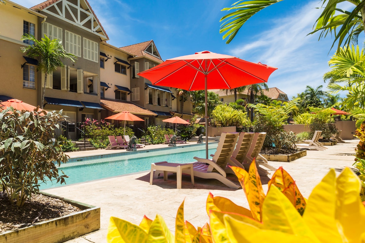 A pool area features a shimmering blue swimming pool, surrounded by tropical plants and several lounge chairs. Bright red umbrellas provide shade, enhancing the inviting atmosphere. The backdrop reveals well-maintained resort buildings under clear blue skies.