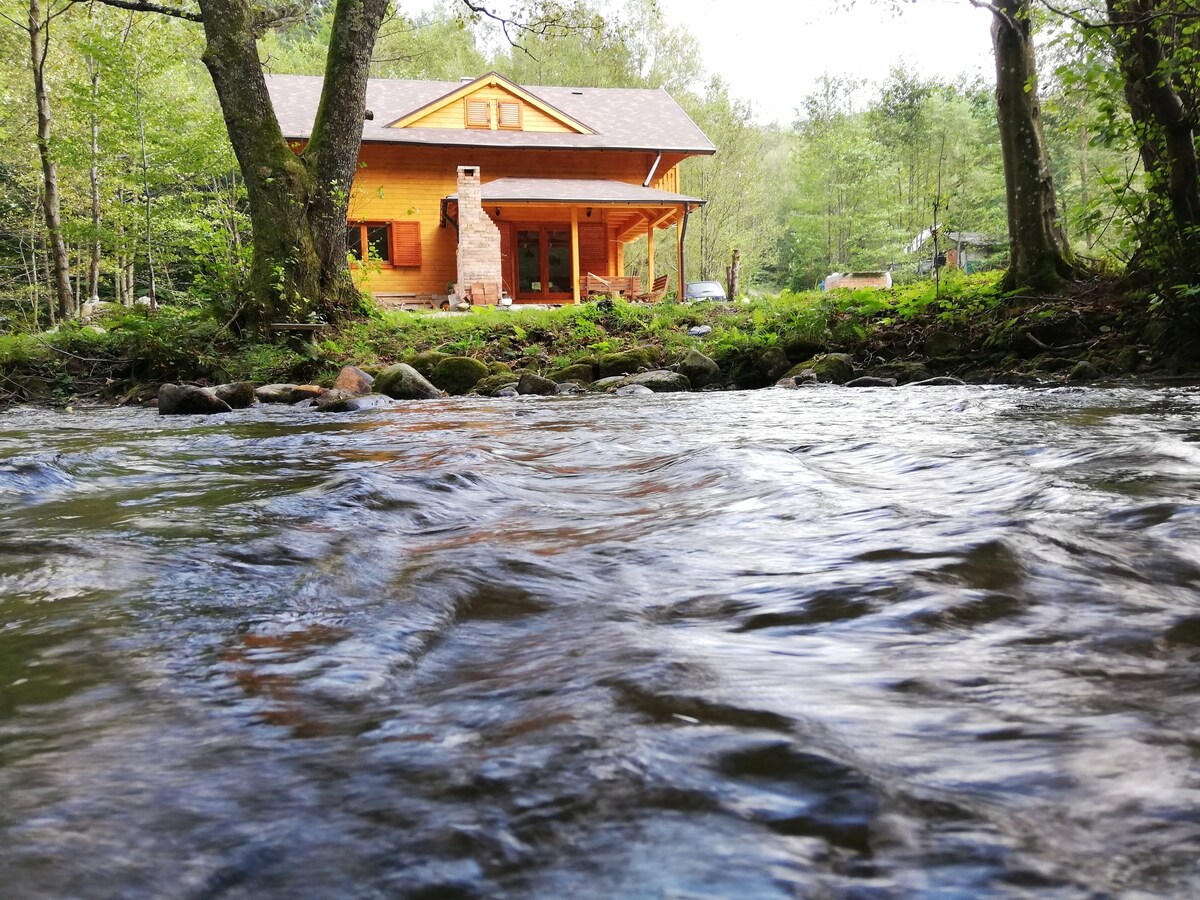 The wooden house is situated beside the flowing Barzava river, framed by lush greenery. A stone chimney adds rustic charm, while the porch invites relaxation with its scenic views of the surrounding nature.