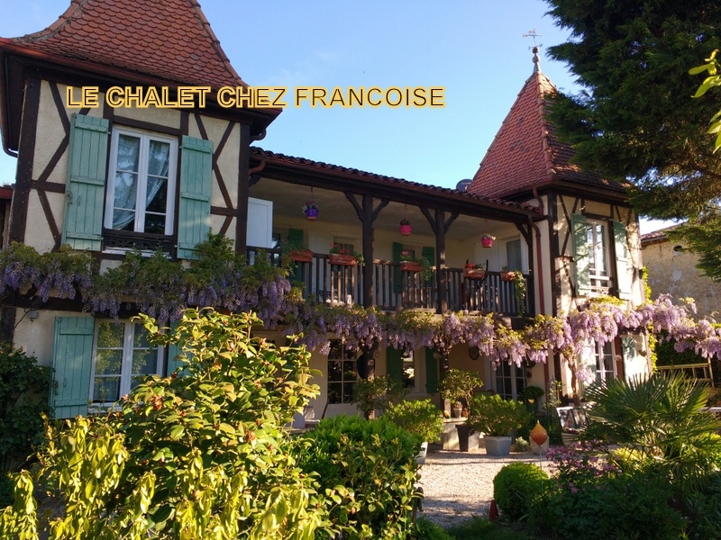 The exterior of the chalet is showcased, featuring two stories with traditional architecture, distinct gabled roofs, and light blue shutters. A balcony adorned with flower pots stretches across the front, surrounded by greenery and colorful plants, creating a welcoming facade.