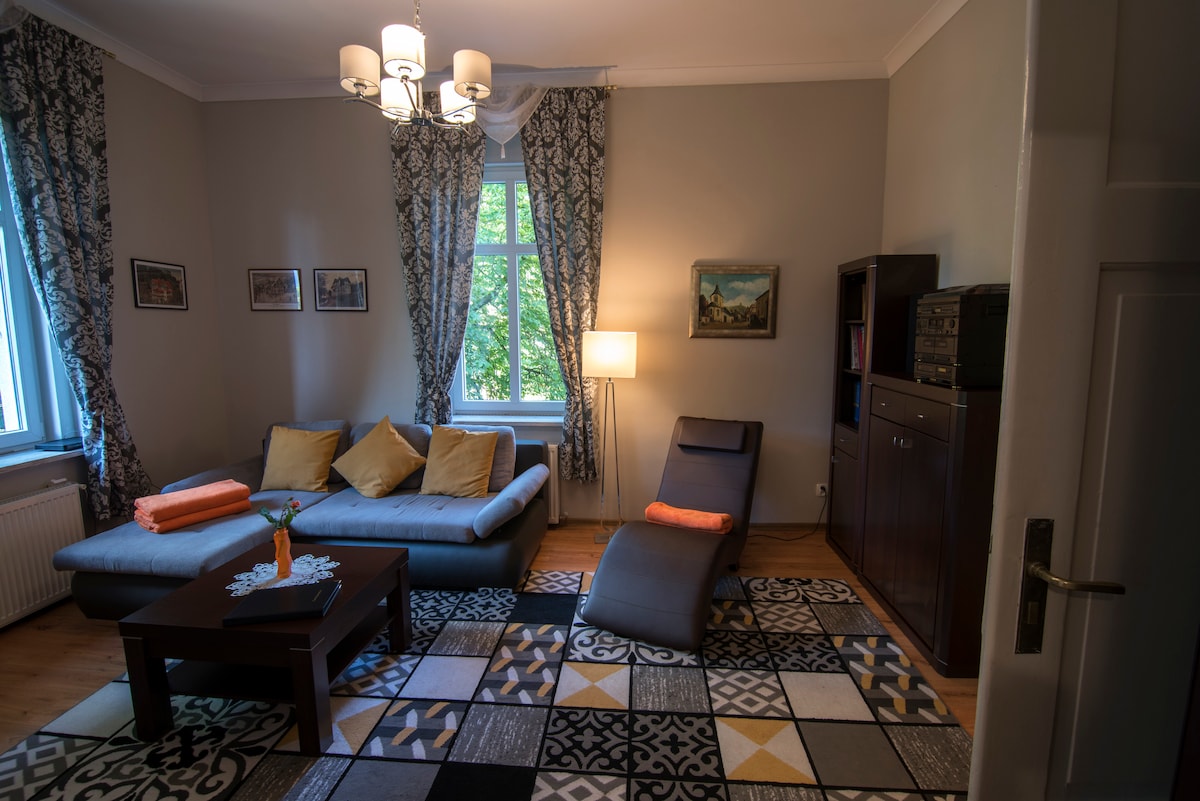 A spacious living area features a gray sofa adorned with orange cushions. A cozy armchair rests next to a floor lamp, illuminating the room. Large windows are framed by patterned curtains, allowing natural light to fill the space. A decorative rug adds a touch of color to the wooden flooring.