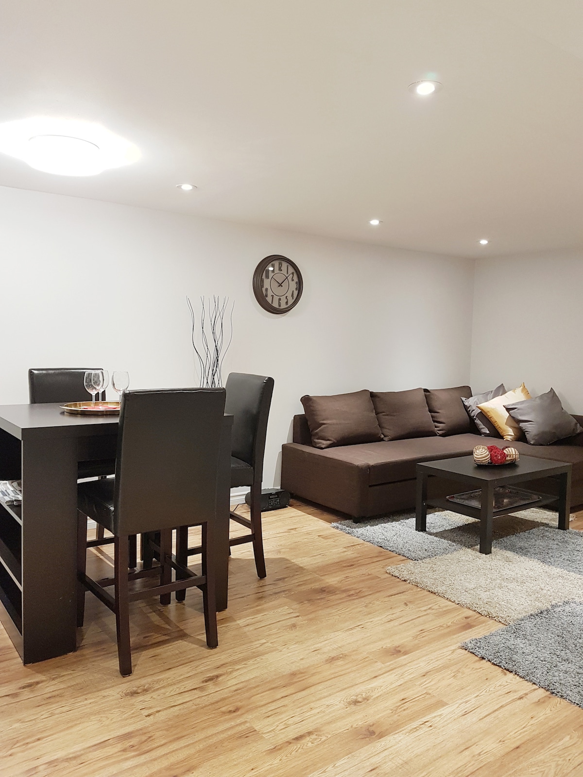 A simple living and dining area is presented, featuring a dark wooden table with four chairs. A comfortable sofa, adorned with decorative cushions, sits next to a low coffee table. Soft lighting is provided by recessed ceiling lights, and a wall clock adds a practical detail.