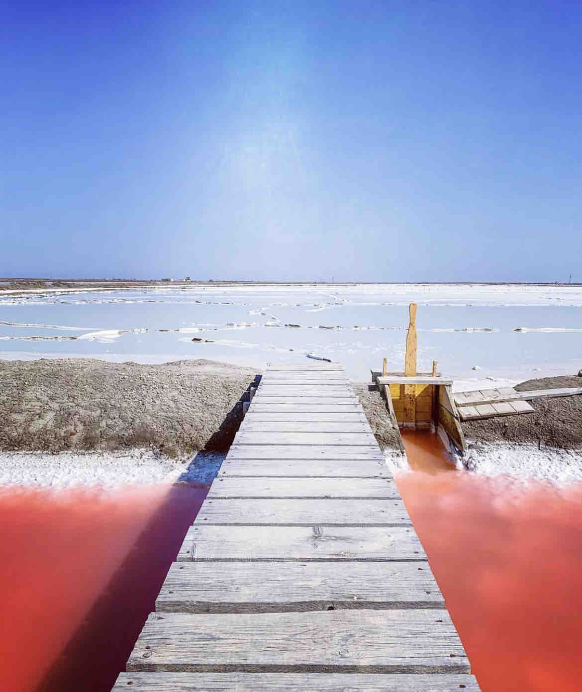A wooden dock extends over a body of water with vibrant red tones, leading towards a flat, salt-covered landscape under a clear blue sky. The serene scene showcases the interplay of colors between the water and the saline deposits.