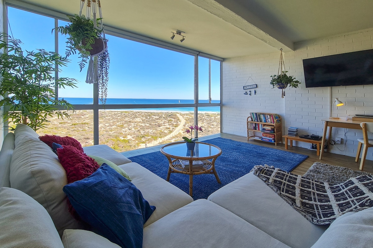 A spacious lounge area features a comfortable couch adorned with cushions. Floor-to-ceiling windows offer an unobstructed view of the coastline. A coffee table and bookshelf are included, complemented by potted plants and a flat-screen TV mounted on the wall.