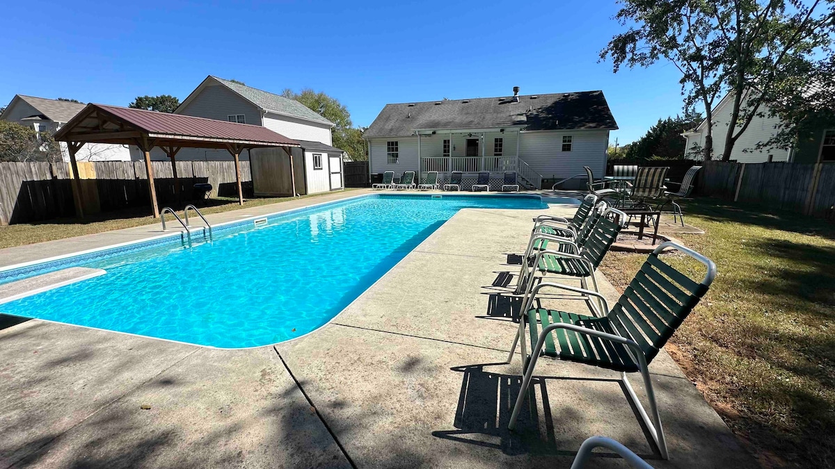 A spacious backyard showcases a large swimming pool, with a shallow end and diving board visible. Several green lounge chairs line the poolside, and a shaded pavilion is present in the background. The area is surrounded by a fence, providing a private atmosphere.