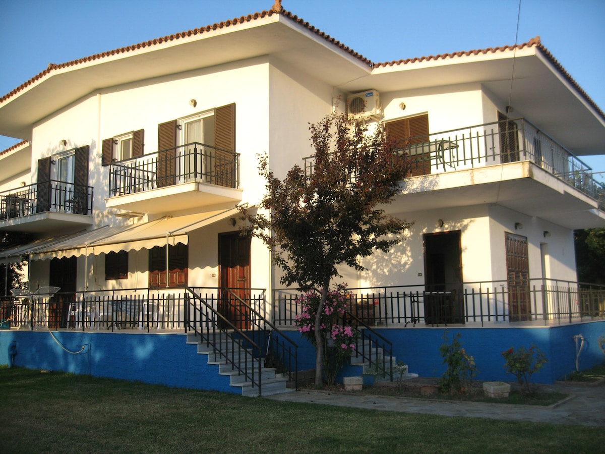 The exterior of the building features a whitewashed façade with dark wooden doors and shutters. Two spacious balconies are visible on the upper floor, surrounded by a lush green lawn and flowering plants. A simple staircase leads up to the entrance.