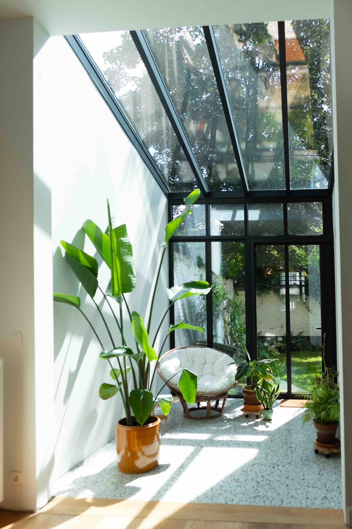 A bright sunroom features large glass panels allowing natural light to fill the space. A plush white chair is positioned beside a tall green plant in a terracotta pot, with smaller plants arranged on the floor, enhancing the serene atmosphere.