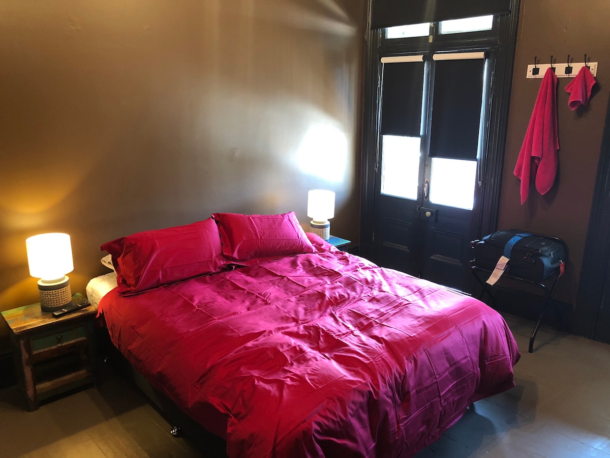 A spacious room features a large bed with a deep red coverlet and matching pillows. Two bedside lamps provide soft lighting. A luggage rack sits at the foot of the bed, while a pair of hangers is mounted on the wall beside a vibrant red towel.