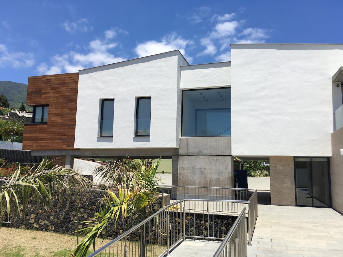 A modern two-story villa features a combination of textured exterior finishes, including smooth white walls and wooden accents. Large windows provide natural light, while a sleek outdoor staircase leads to a landscaped area with greenery. The clear blue sky complements the architectural design.