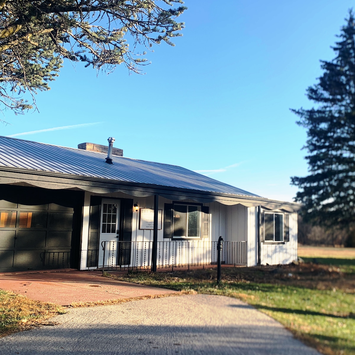 The exterior of the cottage is presented, featuring a metal roof and a covered entrance. Large windows allow natural light to fill the interior, and the inviting pathway is bordered by green grass and trees, enhancing the connection to the surrounding nature.