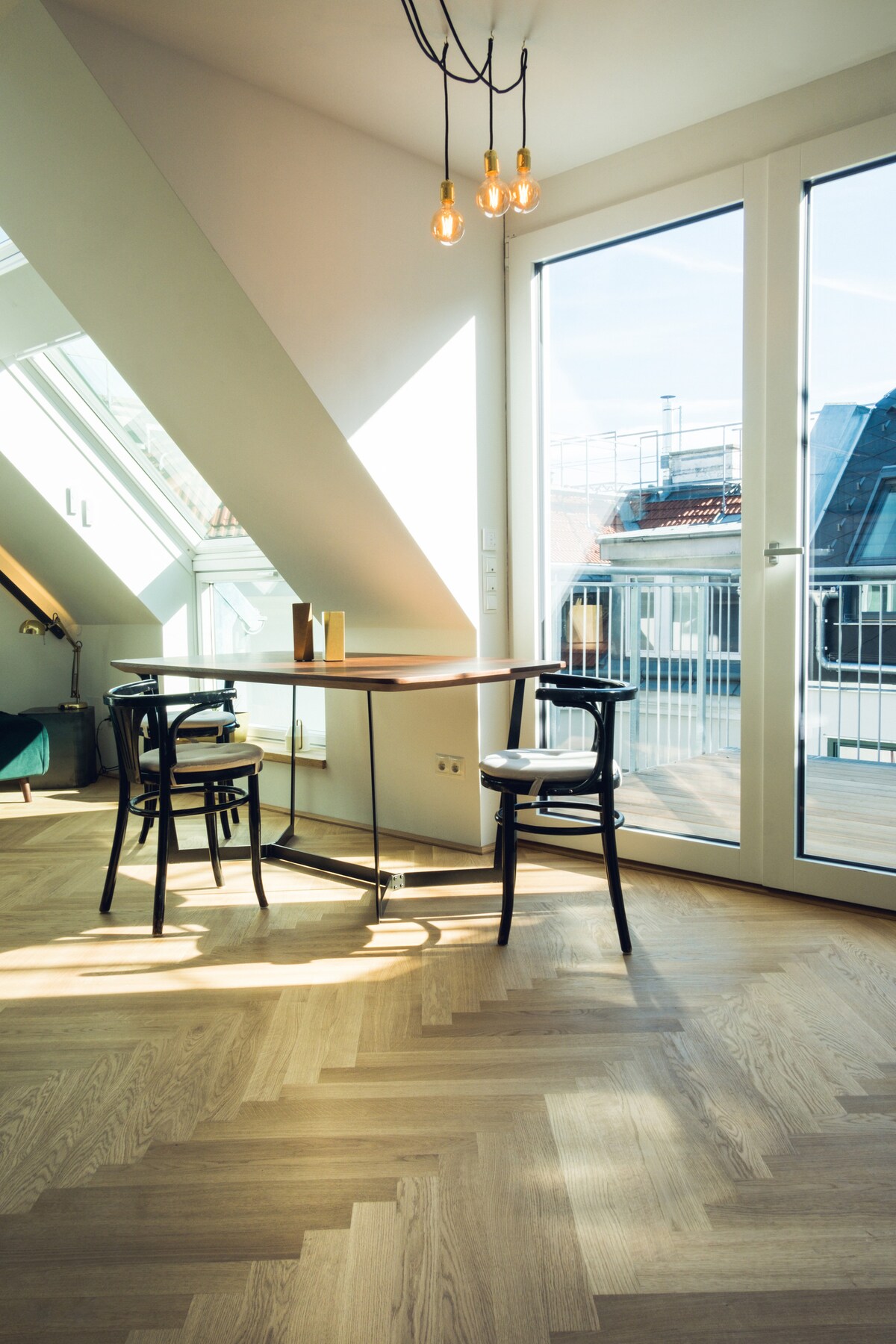 A corner dining area features a round table set against large windows, allowing natural light to fill the space. Two black wooden chairs complement the minimalistic design, while pendant lights hang above, highlighting the warm wood flooring.