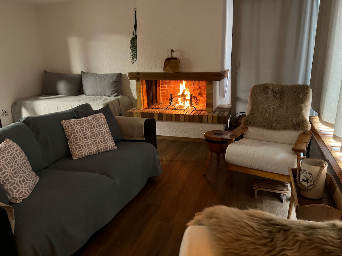 A cozy living area features a fireplace with a warm glow, surrounded by two comfortable sofas and a plush armchair. Natural light enters through the window, and soft blankets are draped over the seating, creating a relaxed atmosphere.