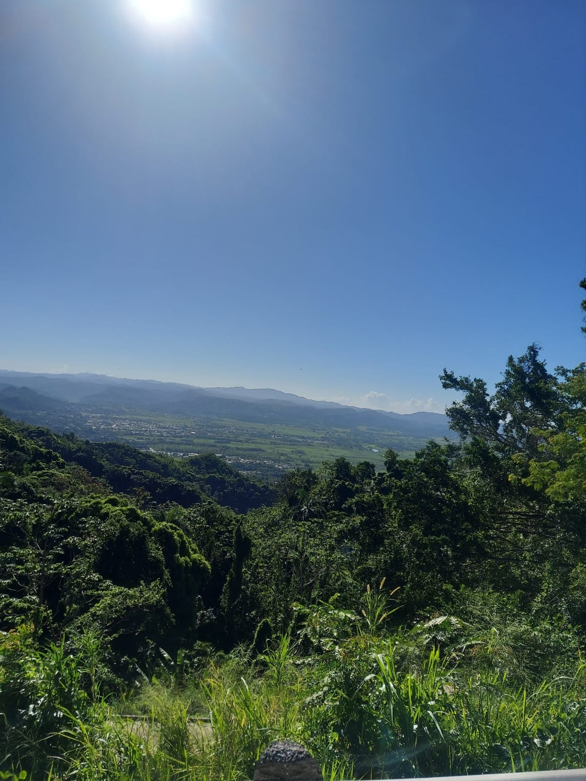 An expansive view of lush green hills is displayed, with a bright sun shining in the clear blue sky. The landscape features a valley below, surrounded by distant mountains, creating a serene natural setting.