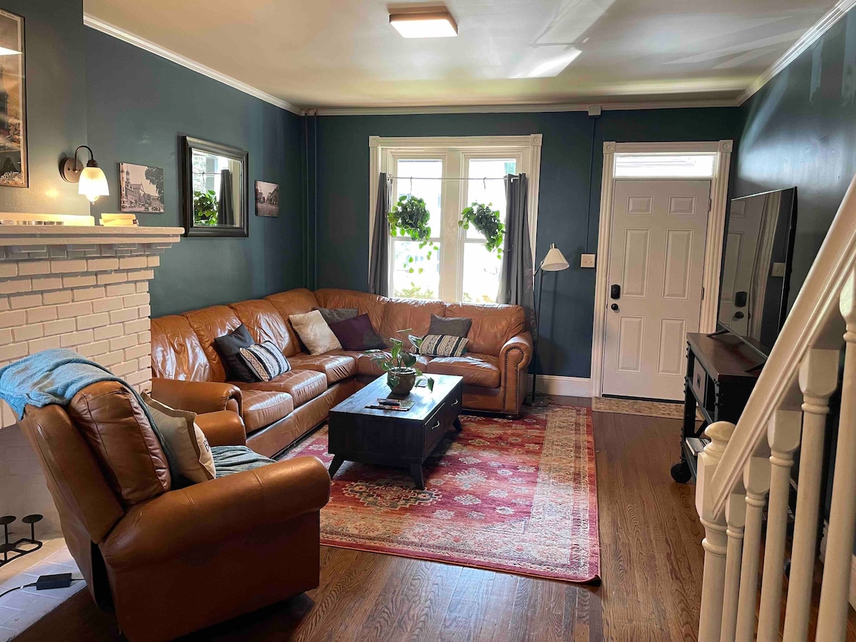 A comfortable living room features a leather sofa and an armchair positioned around a dark wood coffee table. A decorative area rug adds warmth to the hardwood floor. The space is illuminated by natural light through windows adorned with hanging plants, and a staircase is visible in the background.