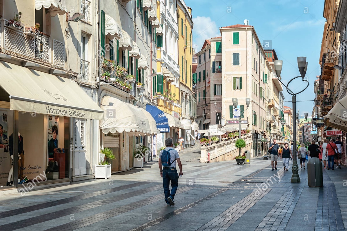 A vibrant street scene is depicted, featuring a pedestrian walking along a wide cobblestone path. Colorful buildings line the street, with outdoor seating and shop awnings visible. Planters filled with greenery add a touch of nature to the urban landscape under a clear blue sky.