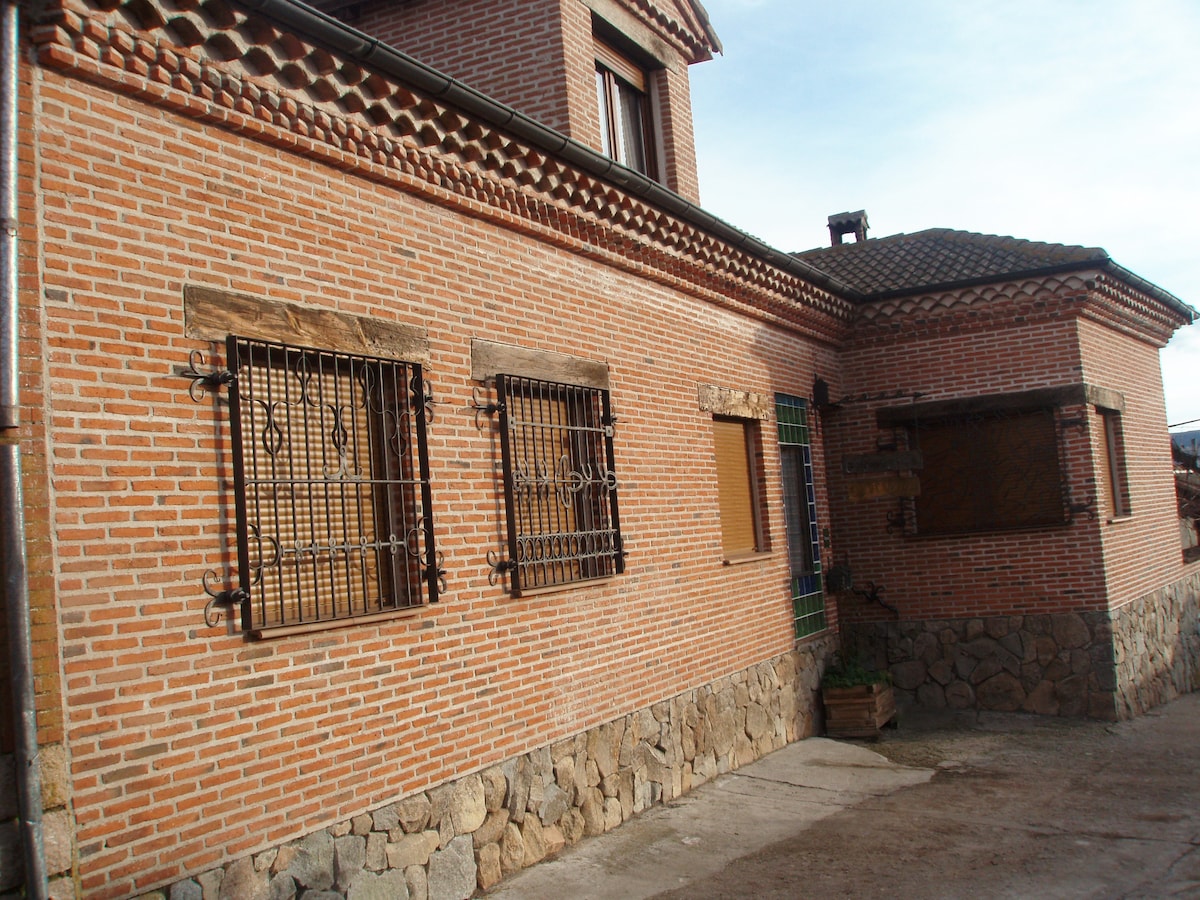The exterior of the house features a combination of red brick and stone, showcasing decorative window bars and wooden window covers. A stone wall serves as a foundation, complementing the structure's rustic charm. The entrance is accentuated by a green door.
