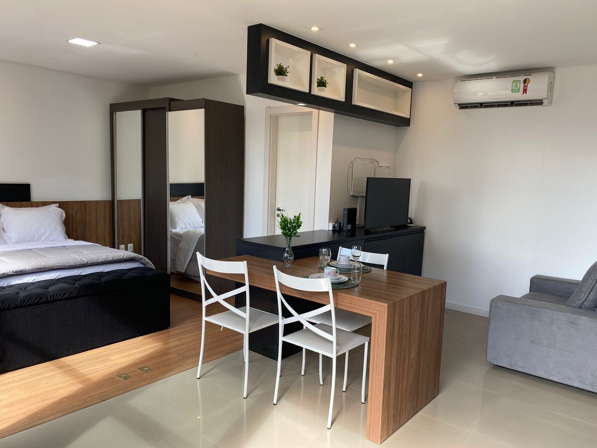 A modern studio space features a cozy sleeping area with a spacious bed and a sleek wardrobe. A dining table for four is placed next to a compact kitchen area, which includes a smart TV and comfortable seating.
