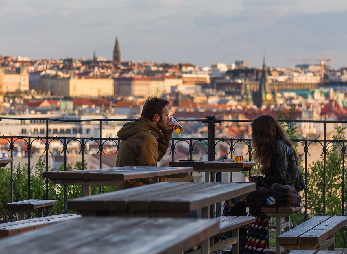 An outdoor seating area features wooden tables and benches, providing a relaxed atmosphere. In the background, a panoramic view of Prague’s skyline is seen, showcasing diverse architectural styles under a golden light. The scene captures a moment of casual conversation with the city as a backdrop.