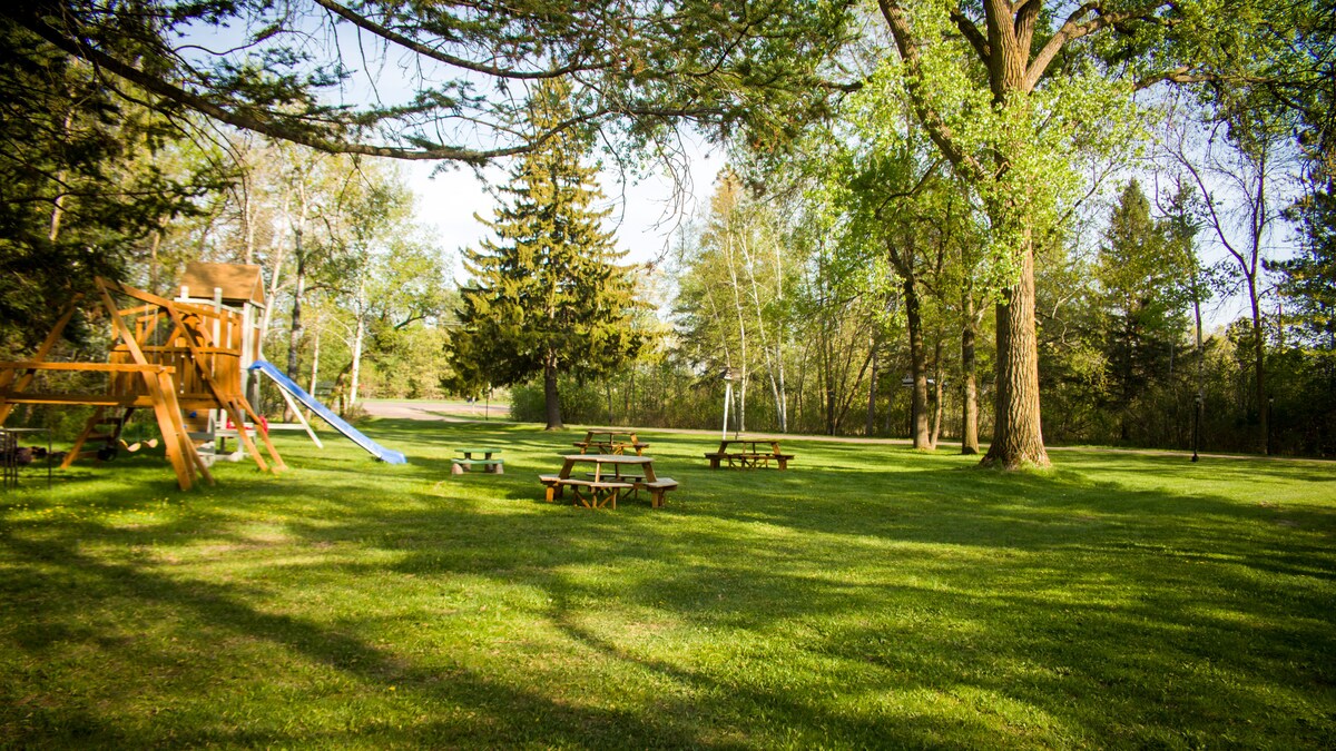 A spacious outdoor area is surrounded by lush greenery, featuring a wooden play structure with a slide. Multiple picnic tables are placed on the well-maintained grass, offering a welcoming space for relaxation and gatherings beneath the shade of tall trees.