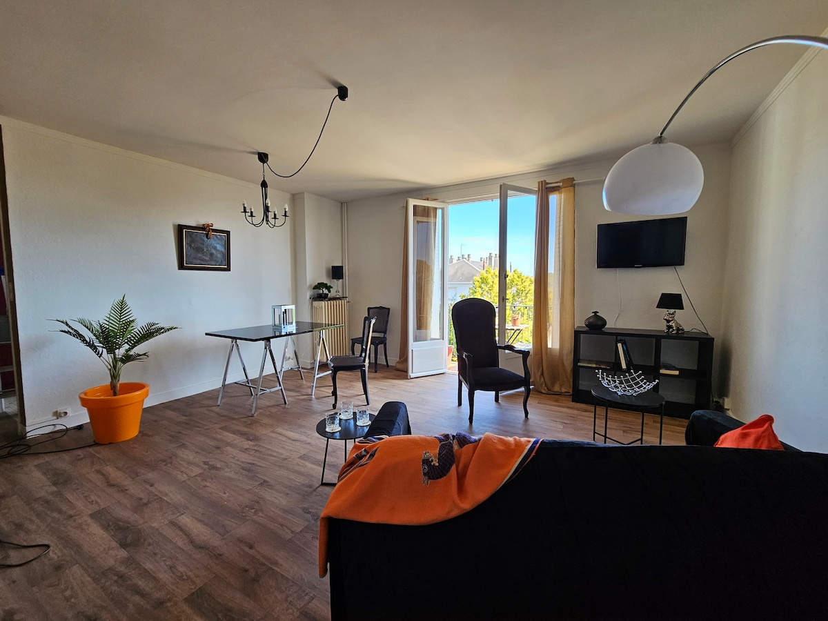 A spacious living area features large windows that allow natural light to fill the room. A comfortable black sofa is complemented by a bright orange throw. A glass table and chairs are positioned near a potted plant, while a screen and decorative elements enhance the setting.
