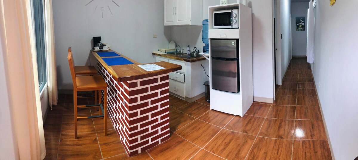 A well-lit kitchen area features a bar with two wooden stools and a countertop made of brick. Appliances include a microwave, a refrigerator, and a coffee maker, arranged along the wall. Smooth wooden flooring continues into a spacious hallway leading to other areas.