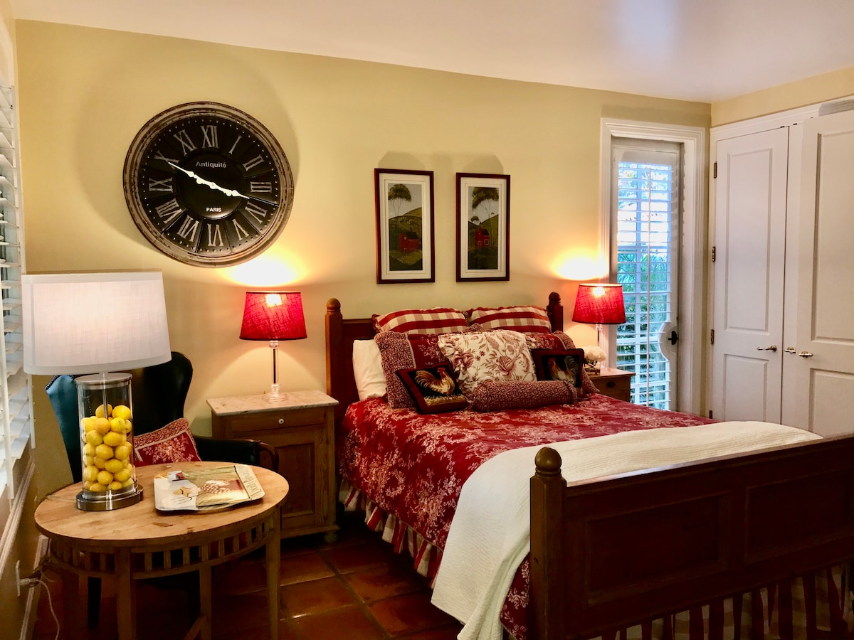 A cozy guest room features a wooden bed adorned with red and white patterned linens. Two bedside lamps cast a warm glow, while a large clock and framed artwork decorate the walls. A small table holds a magazine and a decorative jar filled with lemons.