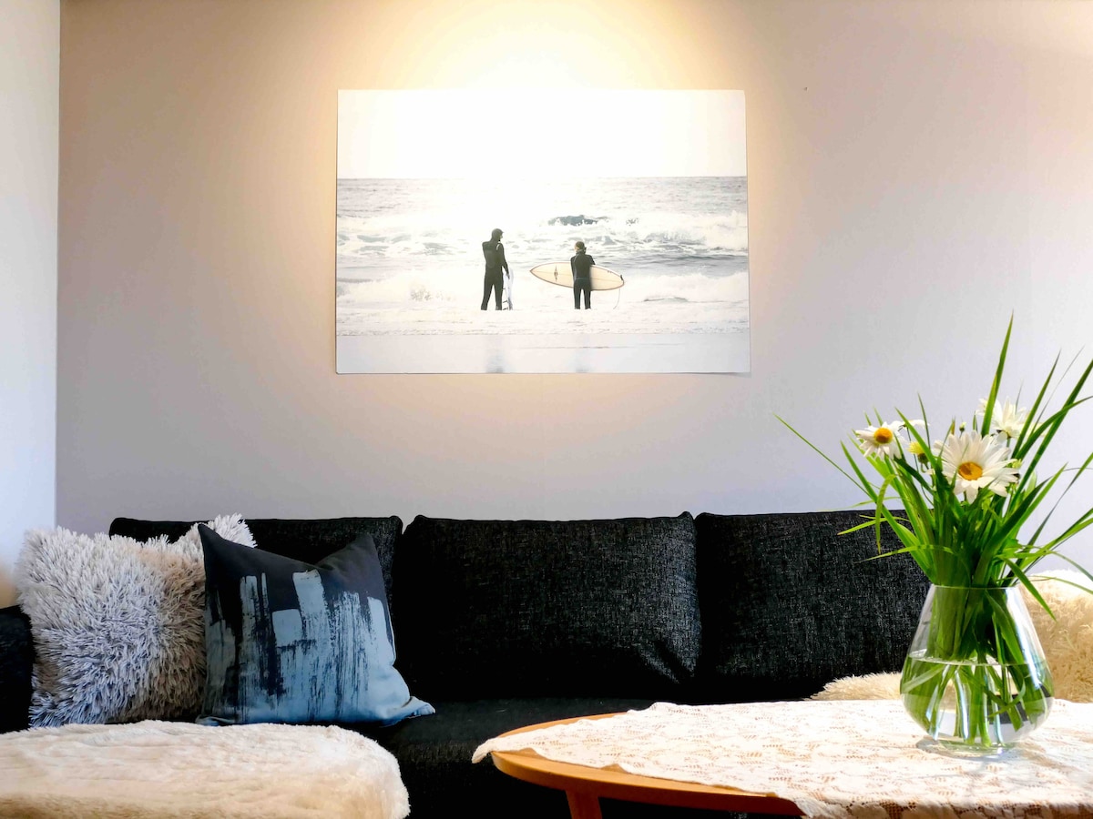A dark sofa is positioned against a light wall, adorned with a large photograph of two surfers by the ocean. A small coffee table features a clear vase filled with fresh flowers, and soft cushions in varying textures are arranged on the sofa.