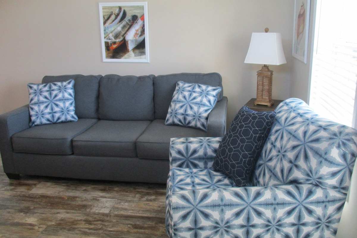 A comfortable seating area features a gray sofa adorned with blue and white patterned throw pillows. Nearby, a patterned armchair complements the sofa. A lamp and wall art enhance the light-filled space, with natural light filtering through white blinds.