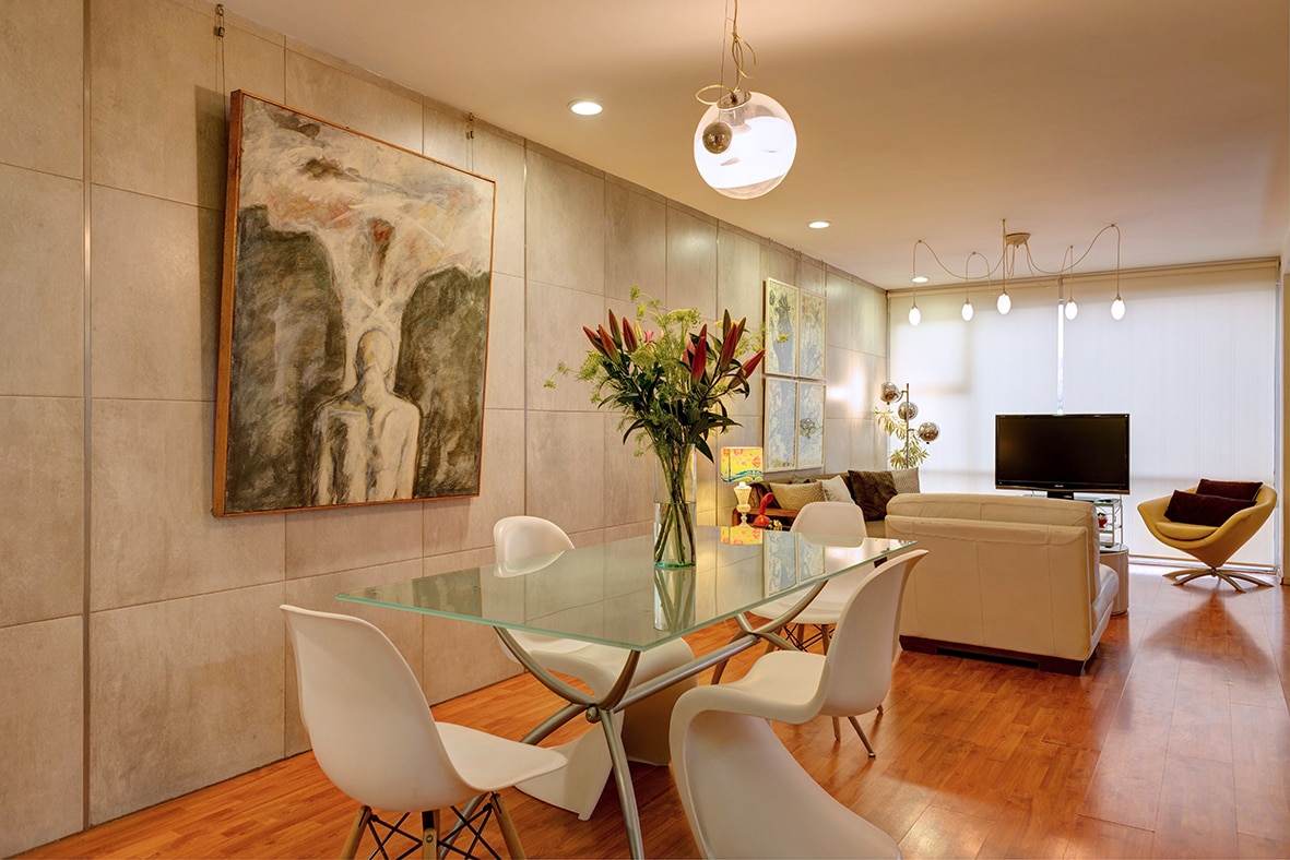 A modern dining area features a glass table surrounded by white chairs, complemented by a large artwork on the wall. Soft lighting is provided by pendant lights above, while a comfortable seating area with a television is visible in the background.