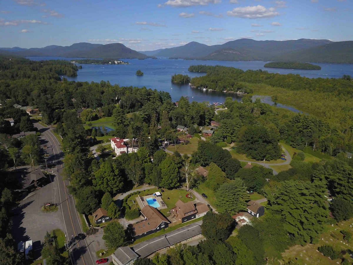 An aerial view captures the scenic landscape of Bolton Landing, featuring a combination of vibrant greenery and the tranquil waters of Huddle Bay. The image showcases a mix of residential structures, a pool area, and natural surroundings, emphasizing the area's accessibility to outdoor activities.