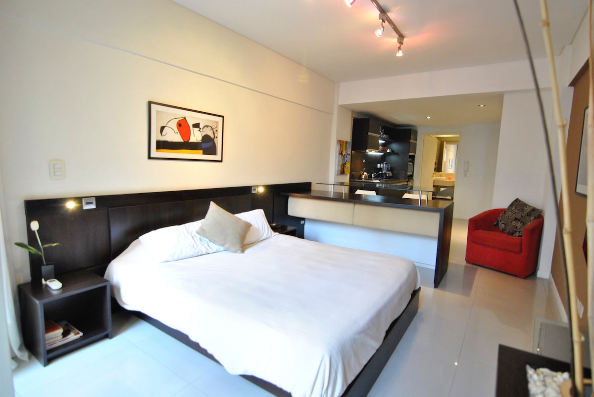 A well-lit studio features a spacious bed with a soft white coverlet, framed by dark wood furniture. A small dining area is visible, along with a modern kitchenette in the background. A pop of color is added by a red armchair in the corner.