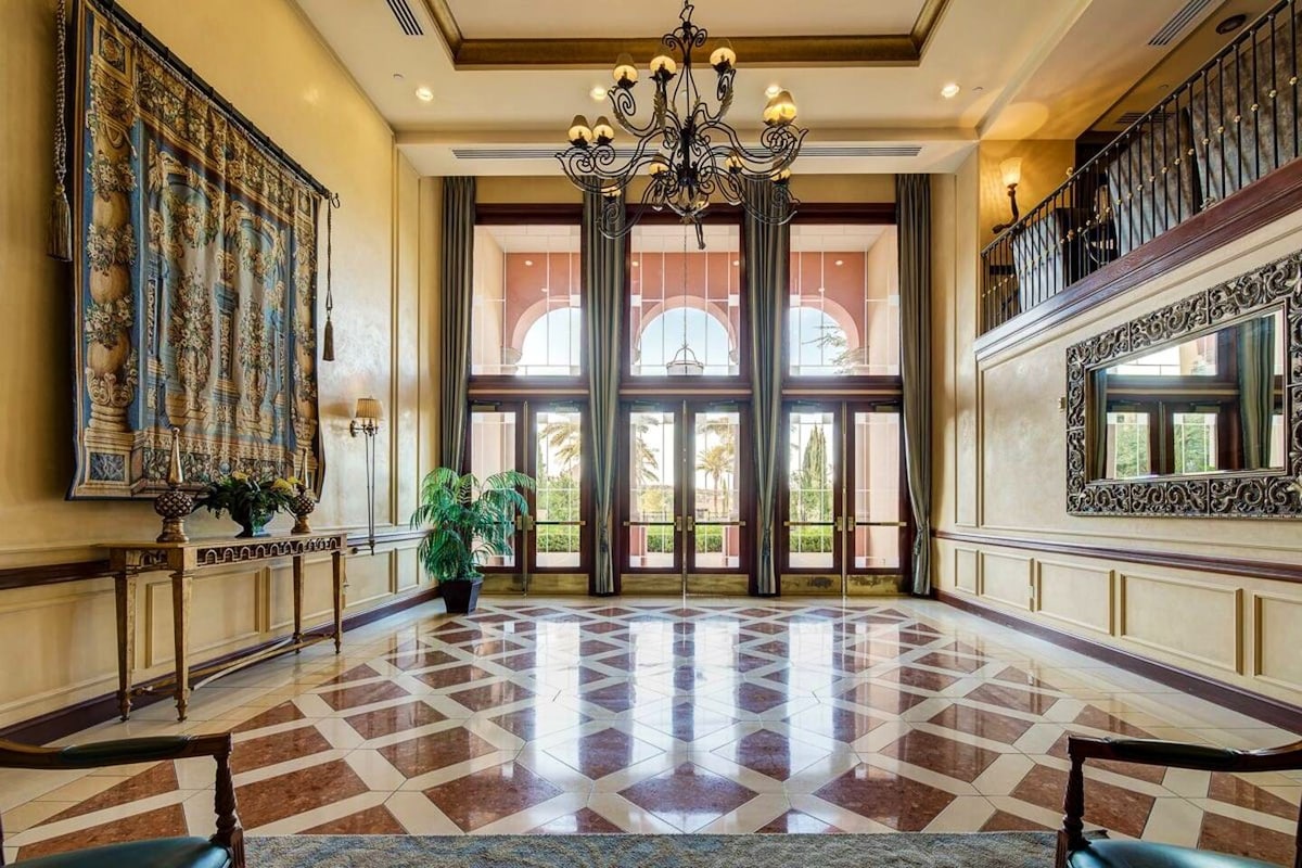 The spacious lobby features elegant marble flooring arranged in a diamond pattern. Large arched windows frame views of the outdoors while decorative chandeliers hang from the ceiling. A grand mirror reflects the sophisticated decor, and lush greenery adds a touch of nature.