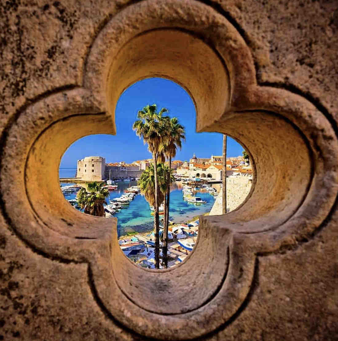 A stone archway, shaped like a flower, frames a vibrant view of the harbor. Tall palm trees sway gently in the foreground, while boats are anchored in the sparkling blue water. The historic buildings of Dubrovnik serve as a picturesque backdrop.