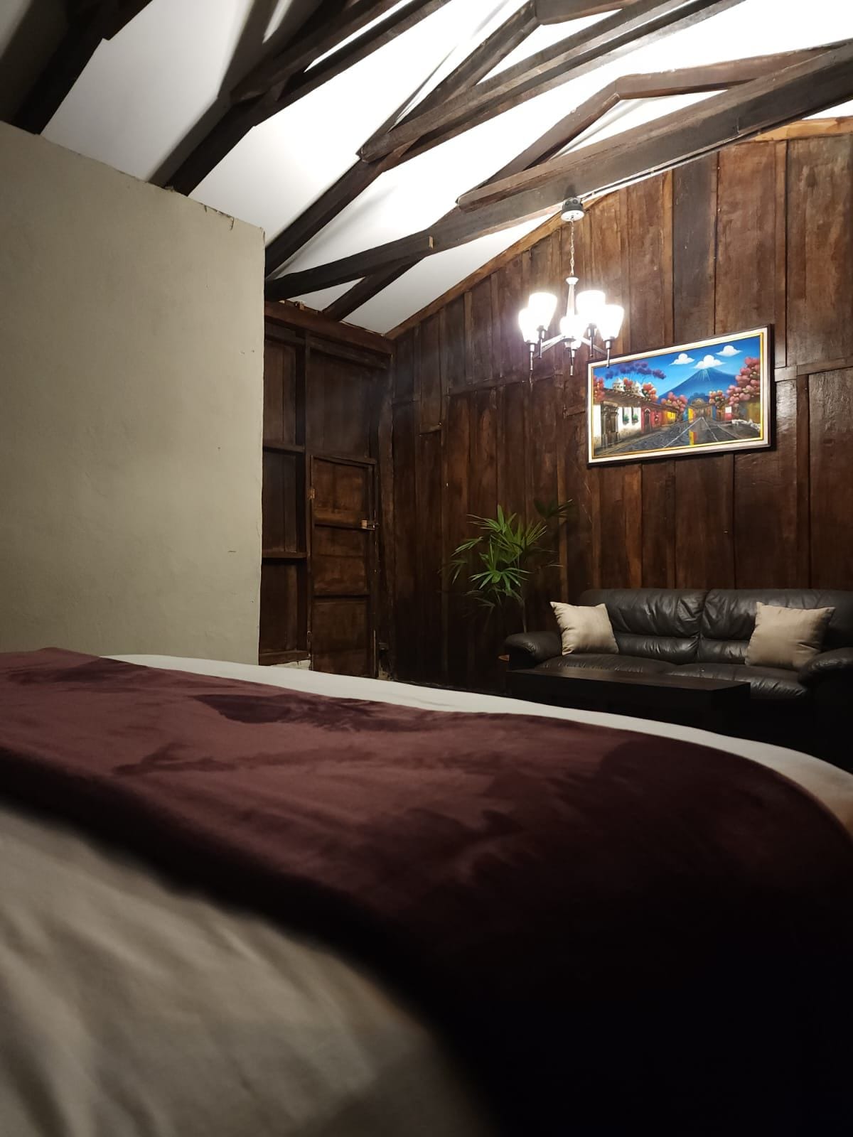 A cozy bedroom is presented with wooden walls and a high ceiling. A dark leather sofa is positioned against the wall, complemented by a colorful framed artwork above it. Soft lighting from a chandelier creates a warm ambiance.