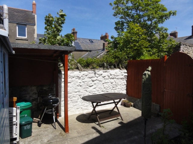 An outdoor space is featured with a wooden table set on a paved surface. A barbecue grill and green recycling bins are also present. A white stone wall borders the area, complemented by green foliage and a wooden gate, creating a private atmosphere.