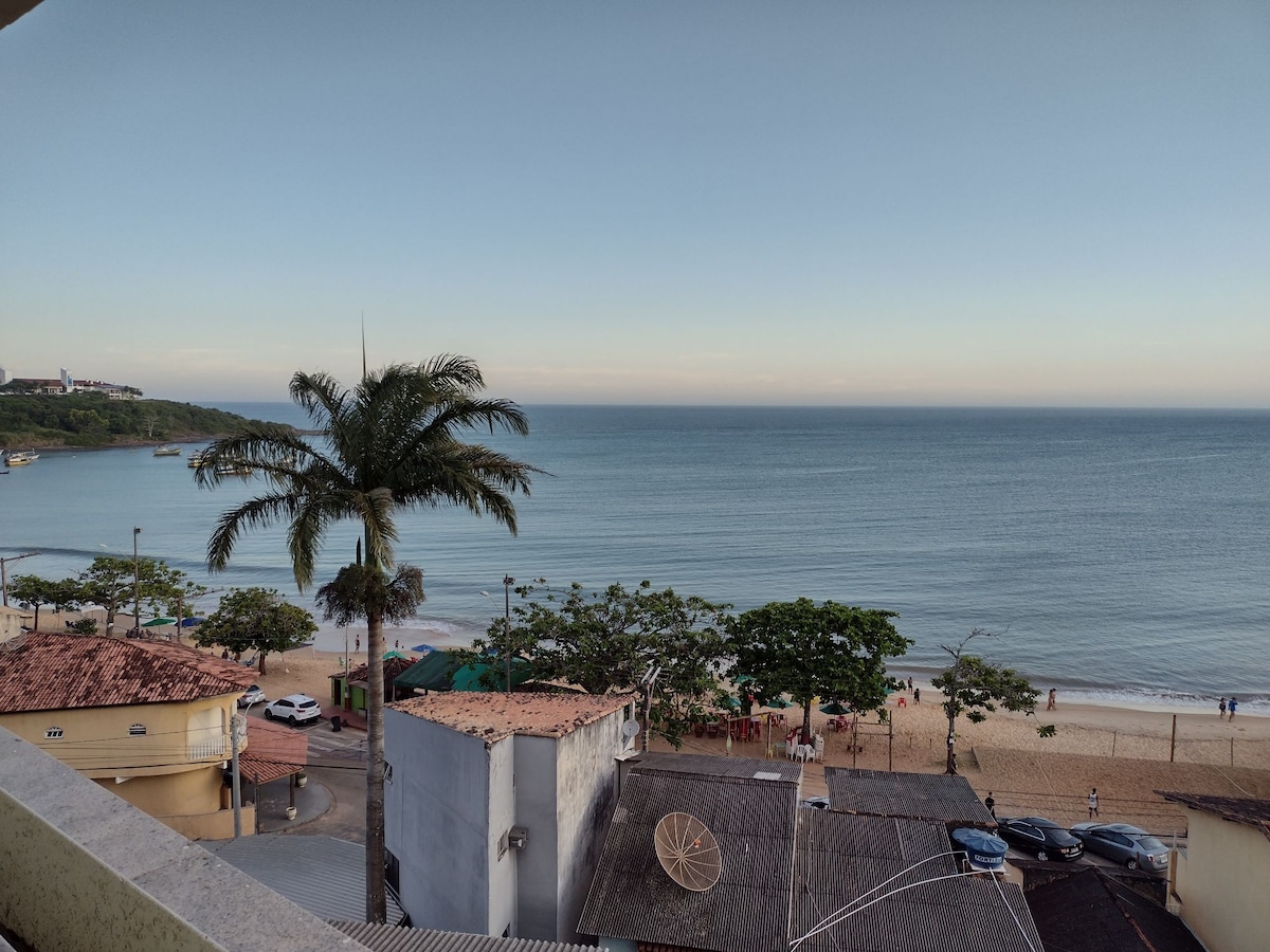 A panoramic view of the beach is captured, showcasing calm waters meeting a gently sloping shoreline. Lush trees line the beach area, while rooftops and parked cars are visible in the foreground. The sky presents a soft gradient as daylight transitions.