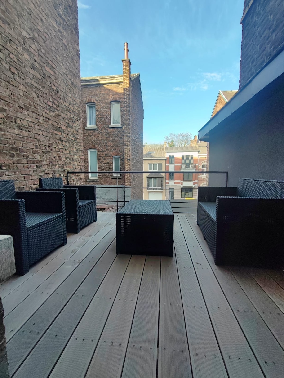 A spacious outdoor terrace is furnished with several dark wicker seating elements, positioned on a wooden deck. The surrounding brick walls offer a sense of privacy while allowing natural light. The sky above is clear, creating an inviting atmosphere for relaxation.