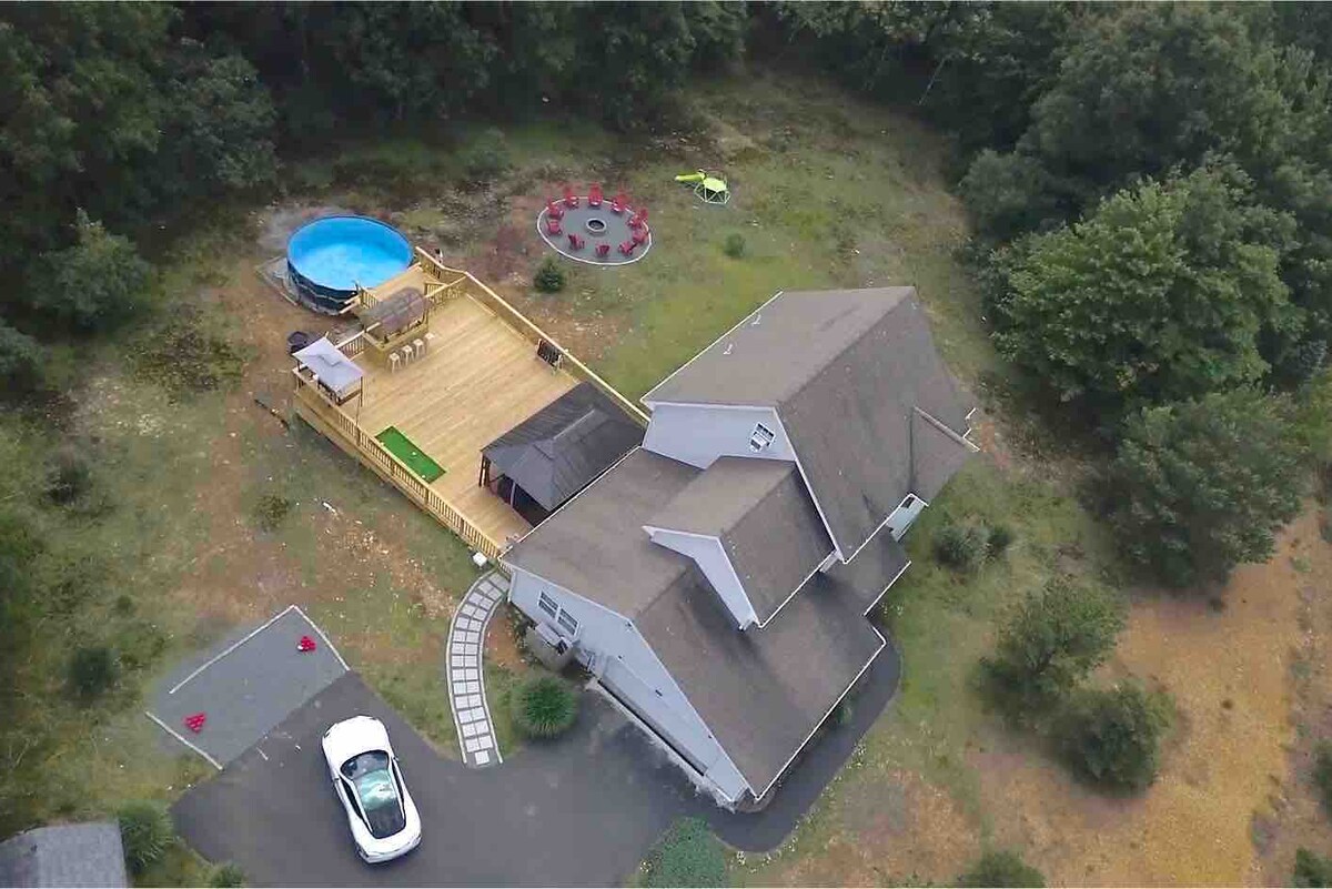 An aerial view of a spacious two-story home surrounded by greenery. A large deck features a circular swimming pool and a fire pit with seating. A driveway is visible, leading to the house, with a lawn area extending into the forested surroundings.