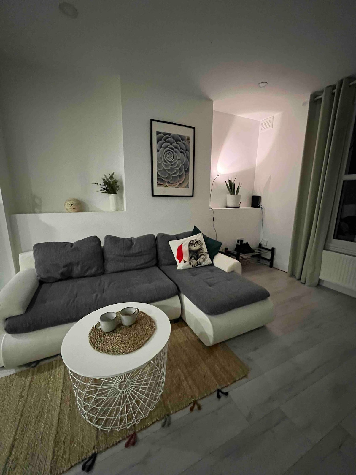 A cozy living space is featured, showcasing a sectional sofa with gray upholstery and a decorative pillow. A round white coffee table sits at the center, accompanied by textured coasters. Soft lighting is provided by a lamp, and a small plant adds a touch of greenery.