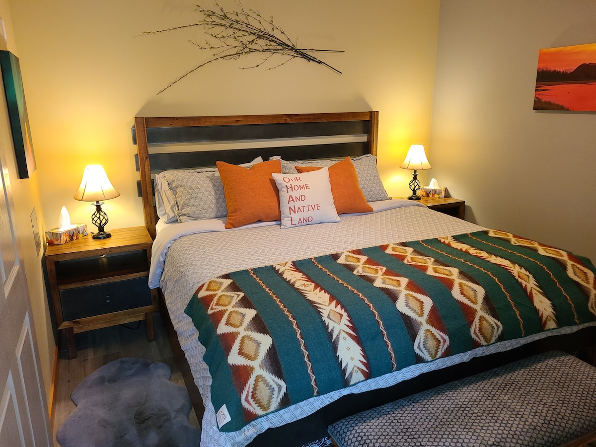 A cozy bedroom features a spacious bed adorned with a patterned blanket and decorative pillows. Soft lighting is provided by two bedside lamps. A rustic wooden headboard and accent wall art enhance the warm atmosphere. A plush rug lies beneath the bed.