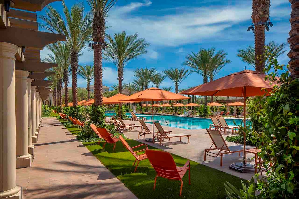 A vibrant outdoor pool area is surrounded by tall palm trees. Orange umbrellas provide shade over lounge chairs arranged on both sides of the pool, with manicured grass lining the pathway. The clear sky enhances the inviting atmosphere of the space.