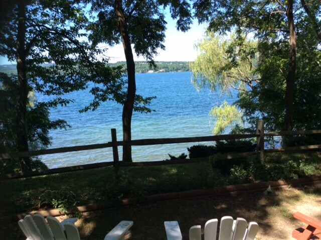 A serene view of the lake is framed by lush greenery and trees. A wooden fence lines the property, providing a sense of separation from the water's edge. Two white adirondack chairs offer a relaxing spot to enjoy the picturesque setting.