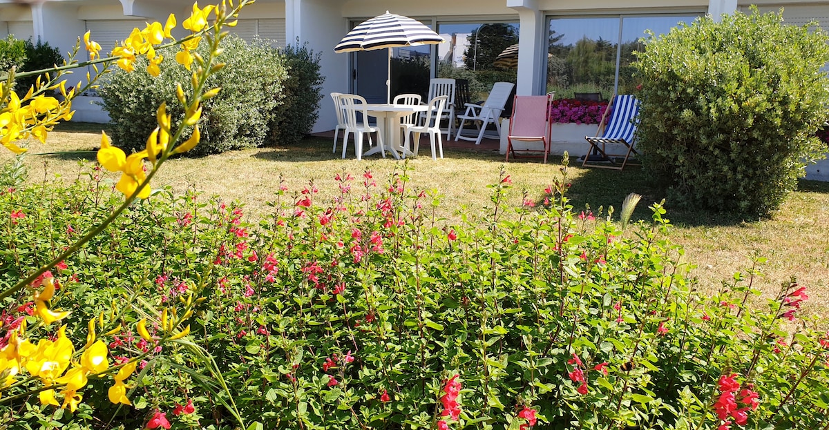 A well-maintained garden area is visible, featuring vibrant flowers in shades of yellow and pink. A shaded outdoor seating arrangement is presented, including a round table and six chairs under a striped umbrella, surrounded by lush greenery and colorful blooms.