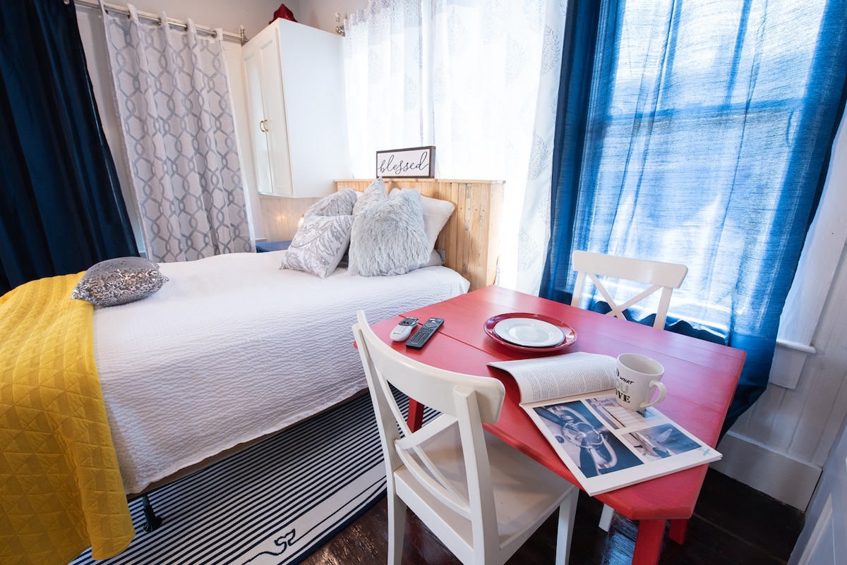 A bright studio features a spacious bed adorned with plush pillows and a colorful throw. A small red dining table is set for two, accompanied by a cozy white chair. Light filters through sheer curtains, creating a warm and inviting atmosphere.